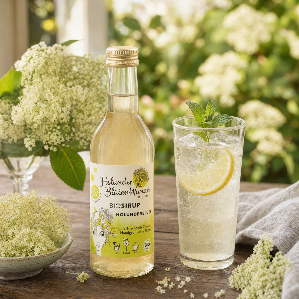 HolunderBlütenWunder Bio-Sirup Holunderblüte mit einem Glas Holunderblütenschorle, Zitrone, Minze und Holunderblüten im Garten